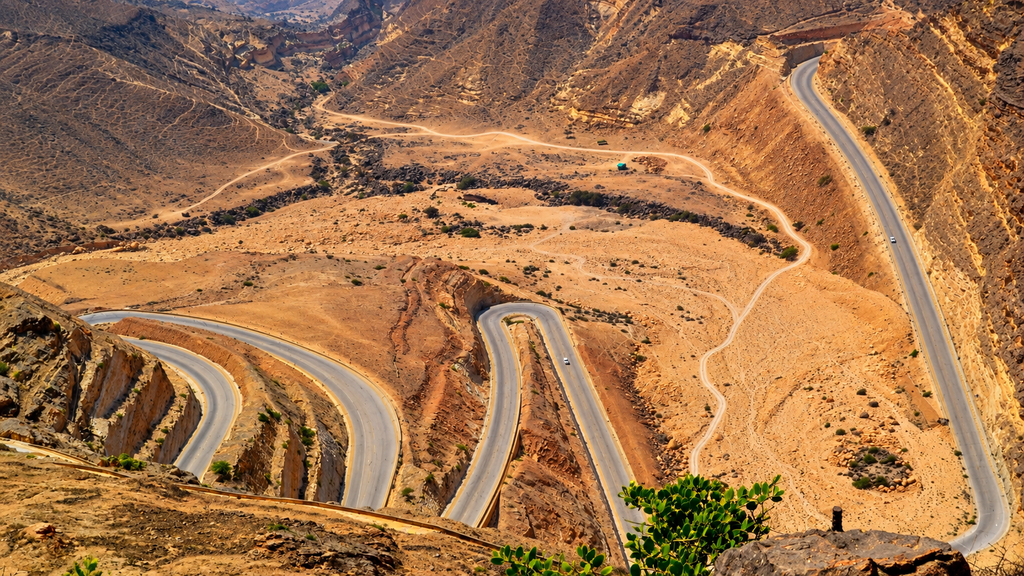 Winding mountain road through the rugged rocky landscape of Oman, viewed from above in warm golden light.
